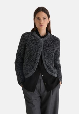 Mujer con cabello oscuro que lleva un cárdigan gris con estampado sobre una camisa negra abotonada y pantalones grises, de pie con las manos en los bolsillos.