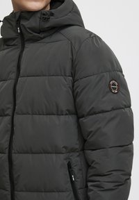 Tamnosivi puffer jackets s kvadratnim uzorkom, s patentnim zatvaračem sprijeda i podesivom kapuljačom. Ima oznaku na lijevoj ruci.