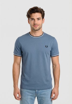 Jonge man met bruin haar, gekleed in een blauw T-shirt met korte mouwen en gestreepte rand, en lichtblauwe jeans, staand tegen een effen achtergrond.