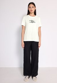 T-shirt vert clair avec le texte noir "evolve-don't compete" sur le devant, associé à un pantalon noir ample et des baskets beiges. Look simple et décontracté.