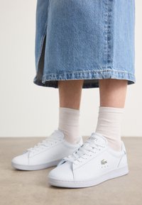 Adidași albi din piele cu o textură netedă, având un logo aurit "Lacoste" și perforații discrete, asortați cu șosete în nuanțe deschise și o fustă din denim.