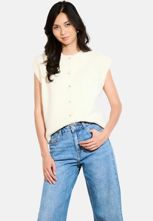 Crème-kleurige mouwloze gebreide top met een ronde hals en knoopsluiting, gecombineerd met lichtblauwe high-waisted jeans.