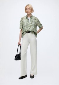 Woman with short blonde hair wearing a green floral short-sleeve shirt, white pants, black shoes, and holding a black handbag.