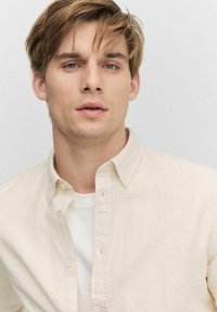 Licht beige button-up shirt over een wit T-shirt, met een kraag en subtiele textuur. Het onderwerp heeft kort, rommelig haar en een neutrale achtergrond.