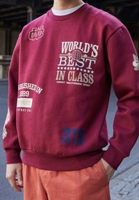 Felpa color marrone con stampe bianche e dorate, inclusa la scritta "WORLD'S BEST IN CLASS" e numeri importanti. Polsini e scollo a coste.