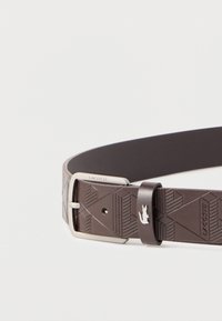 Καφέ δερμάτινη ζώνη με γεωμετρικό ανάγλυφο σχέδιο, ασημένια αγκράφα με χαραγμένο το όνομα της μάρκας και μικρό ασημένιο λογότυπο κροκοδείλου στο θηλάκι.