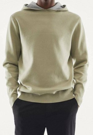 Homme portant un sweat à capuche vert olive clair et un pantalon noir, mains dans les poches, debout devant un fond clair uni.