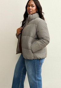 Giubbotto puffo con motivo a scacchi marrone e crema, colletto alto, chiusura con zip e tasche laterali, abbinato a jeans a zampa d'elefante blu.