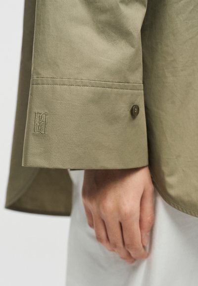 Κοντινό πλάνο μιας μανσέτας πουκαμίσου σε μπεζ χρώμα με κουμπί, κεντημένο λογότυπο και ένα χέρι που ακουμπά δίπλα της πάνω σε ουδέτερο φόντο.