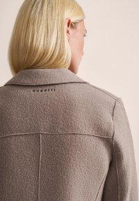 Hellbrauner Wollmantel mit strukturiertem Finish, ausgestattet mit einem Kragen und sichtbaren Nähten. Logo an der Rückseite des Halsausschnitts platziert.