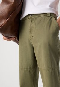Olive green pants with a relaxed fit, featuring side pockets, a smooth texture, and stitched accents. A brown leather bag is held nearby.
