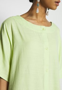 Donna che indossa una camicia verde chiaro con bottoni e lunghe orecchini pendenti con perle e perline verdi.