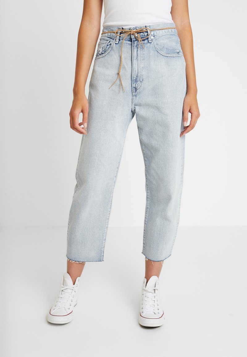 Jeans de mezclilla azul claro con corte holgado, que presentan bolsillos frontales, un dobladillo deshilachado y un cinturón de tela atado. Combinados con zapatillas de lona blancas.