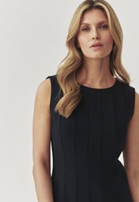 Marineblaues ärmelloses Kleid mit vertikalen Falten, glatter Textur und rundem Ausschnitt. Die Haare sind wellig und fallen über eine Schulter.