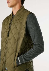 Muž oblečený v olivovo zelenom prešívanom vestu cez tmavošedú dlhú košeľu s nápisom "PEPE JEANS" na manžete, ruka vo vrecku vesty, biely pozadie.
