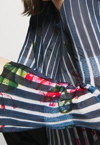 Randig genomskinlig blus med en blandning av mörkblå och vita ränder, prydd med färgglada blommönster i rött och grönt på ärmarna.