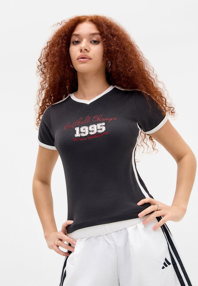 Zwart T-shirt met korte mouwen en V-hals; heeft witte afgewerkte randen, grafisch ontwerp "Softball Champs 1995" in rood en wit, gladden stof, aansluitende pasvorm.