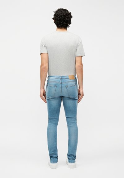 Άτομο με σγουρά σκούρα μαλλιά, που φορά ανοιχτό γκρι κοντομάνικο πουκάμισο, ελαφρώς στενά τζιν ανοιχτού μπλε χρώματος και λευκά αθλητικά παπούτσια, στέκεται με την πλάτη προς τα εμπρός.