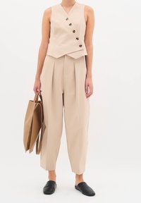 Béžový top bez rukávů s výstřihem do V a pěti tmavými knoflíky, kombinovaný s volnými, plisovanými béžovými kalhotami a černými pantoflemi. Nese hnědou tote kabelku.