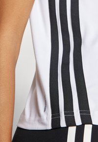 Bílý sportovní top bez rukávů s třemi černými svislými pruhy. Tkanina vypadá hladce a má přiléhavý střih. Viditelná značka "Climacool".