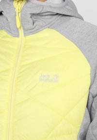 Gul vadderad jacka med grå ärmar, med dragkedjestängning och en logotyp på bröstet. Slät textur med en modern design.