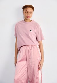 Rosa bomullströja med en liten logopatch på bröstet, tillsammans med rosa satinbyxor med vita sidostråk och en broderad logo.