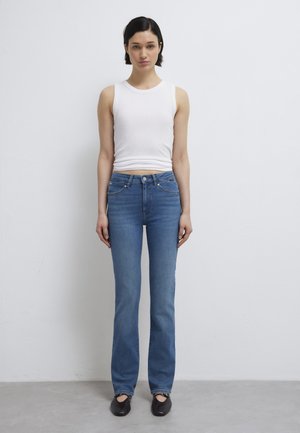 Woman with short dark hair wearing a white sleeveless top, blue jeans, and black shoes, standing against a plain white wall.