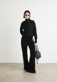 Zwarte wollen coltrui, brede zwarte fluwelen broek met zijstreepjes, zwarte zonnebril en een kleine zwarte handtas. Strakke algehele uitstraling.