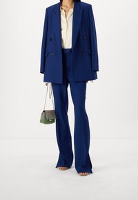 Blå dubbelknäppt kavaj över en krämfärgad blus, ihop med blå utsvängda byxor. En liten grön och silverfärgad handväska hålls i ett kedjerem.