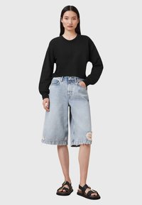 Sudadera corta negra, pantalones cortos de mezclilla desgastados en azul claro con dobladillos deshilachados y sandalias negras con acentos en dorado.