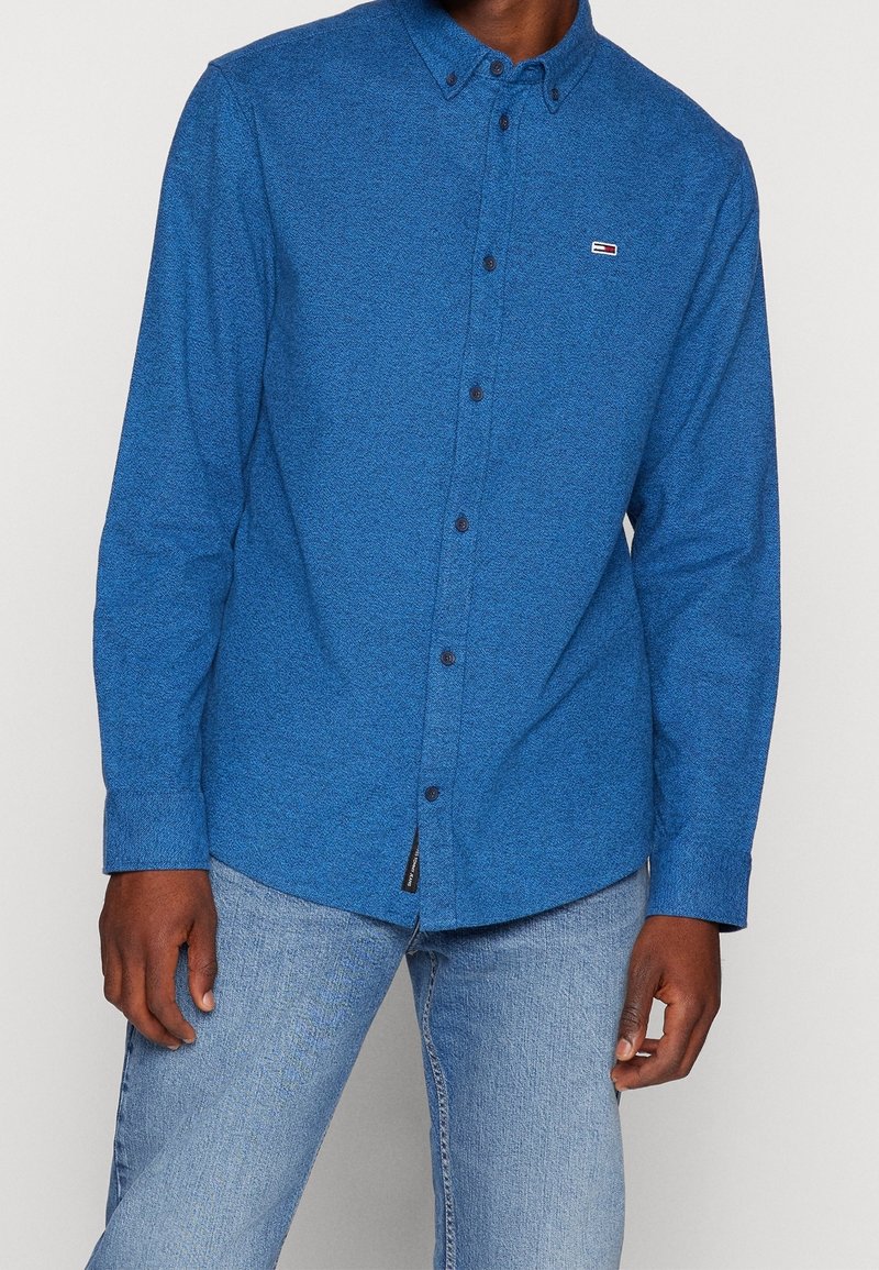 Homme portant une chemise bleue à manches longues avec un petit logo rectangulaire sur la poitrine et un jean bleu clair.