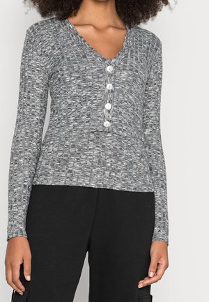 Vrouw met een strak grijs gestructureerd long-sleeve shirt met een V-hals en vier witte knopen aan de voorkant, gecombineerd met zwarte broek.