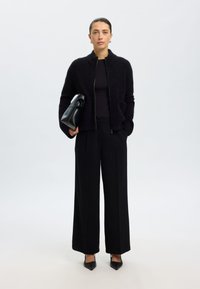 Juoda dirbtinio kailio striukė su užtrauktuku ir aukštu apykakle, derinama su juodu aptemptu viršumi ir plačiomis juodomis kelnėmis. Laiko mažą juodą rankinę.