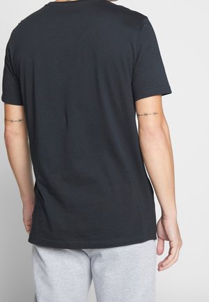 Homme portant un t-shirt noir uni à manches courtes et un pantalon gris clair, vu de dos avec des tatouages de fil barbelé sur les deux bras supérieurs.