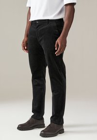 Pantaloni in corduroy neri con una silhouette dritta, caratterizzati da un tessuto testurizzato, tasche frontali e chiusura con bottone. Indossati con scarpe marroni.