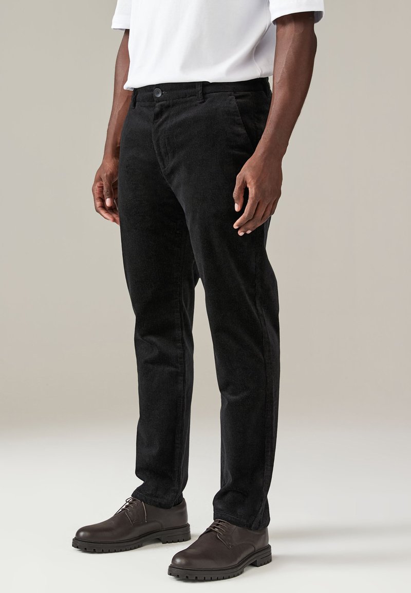Pantaloni in corduroy neri con una silhouette dritta, caratterizzati da un tessuto testurizzato, tasche frontali e chiusura con bottone. Indossati con scarpe marroni.