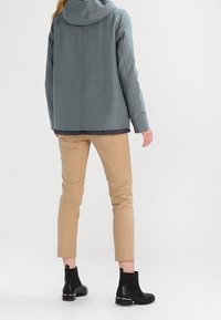 Grå hættejakke med en glat tekstur, beige tilspidsede bukser og sorte ankelstøvler med en skinnende detalje ved hælen.