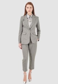 Completo grigio a righe sottili con giacca aderente e pantaloni affusolati. Presenta una camicia grigia chiara sotto. Il modello indossa tacchi argentati.