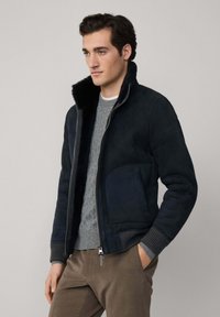 Tamni mornarski sako od nubuk kože s bujnim crnim krznom oko vrata, sa zatvaračem na rajsferšlus i ukrašenim dizajnom, nosi se preko sivo pletenog džempera.