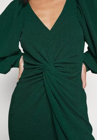 Femme portant une robe texturée vert foncé avec des manches longues bouffantes et un détail de nœud tordu à la taille.