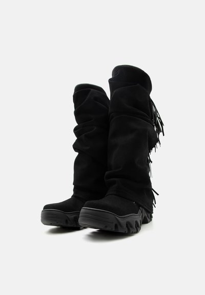 Μαύρες μπότες μέχρι το γόνατο με χοντρές υφασμάτινες σόλες και μαλακό, χαλαρό σουέντ υλικό με φουντίτσες στο πλάι, σε λευκό φόντο.