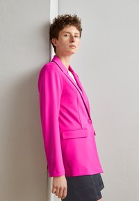 Fuksia blazer med klaffekrage, frontlommer og en enkelt knappelukking. Glatt tekstur og skreddersydd passform, kombinert med et mørkt skjørt.