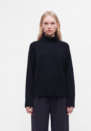 Jeune femme aux longs cheveux blonds porte un pull à col roulé noir et un pantalon large foncé sur un fond blanc uni.