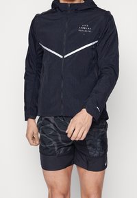 Homme portant une veste de course Nike noire avec des bandes réfléchissantes blanches et un short noir à motifs.
