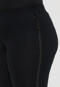 Μαύρες κολάν που διαθέτουν λεία ύφασμα με μια κομψή, γυαλιστερή μαύρη λωρίδα από παγιέτες κατά μήκος της εξωτερικής πλευράς. Στενή σχεδίαση που αναδεικνύει το σχήμα.