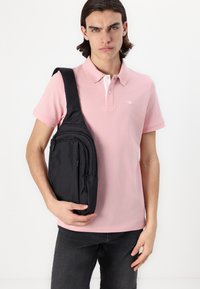 Sac banane noir avec plusieurs compartiments zippés, texture lisse et bandoulière réglable, tenu par une personne portant un polo rose.