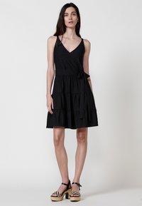 Schwarzes ärmelloses Kleid mit V-Ausschnitt, gestuften Design, strukturiertem Stoff und seitlicher Bindung in der Taille. Kombiniert mit Plateauschuhen.