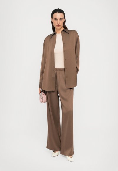 Person wearing loose brown button-up shirt and matching wide-leg pants, white top, white pointed shoes, holding small pink bag.