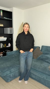 Γυναίκα με μαύρη φούτερ με κουκούλα και μπλε τζιν στέκεται με τα χέρια στις τσέπες δίπλα σε τιρκουάζ καναπέ και μαύρη βιβλιοθήκη σε σαλόνι.