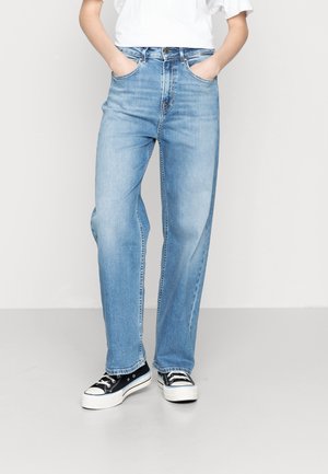 Person wearing light blue straight-leg jeans, white short-sleeve top, and black low-top sneakers with white soles, standing with hands in pockets.